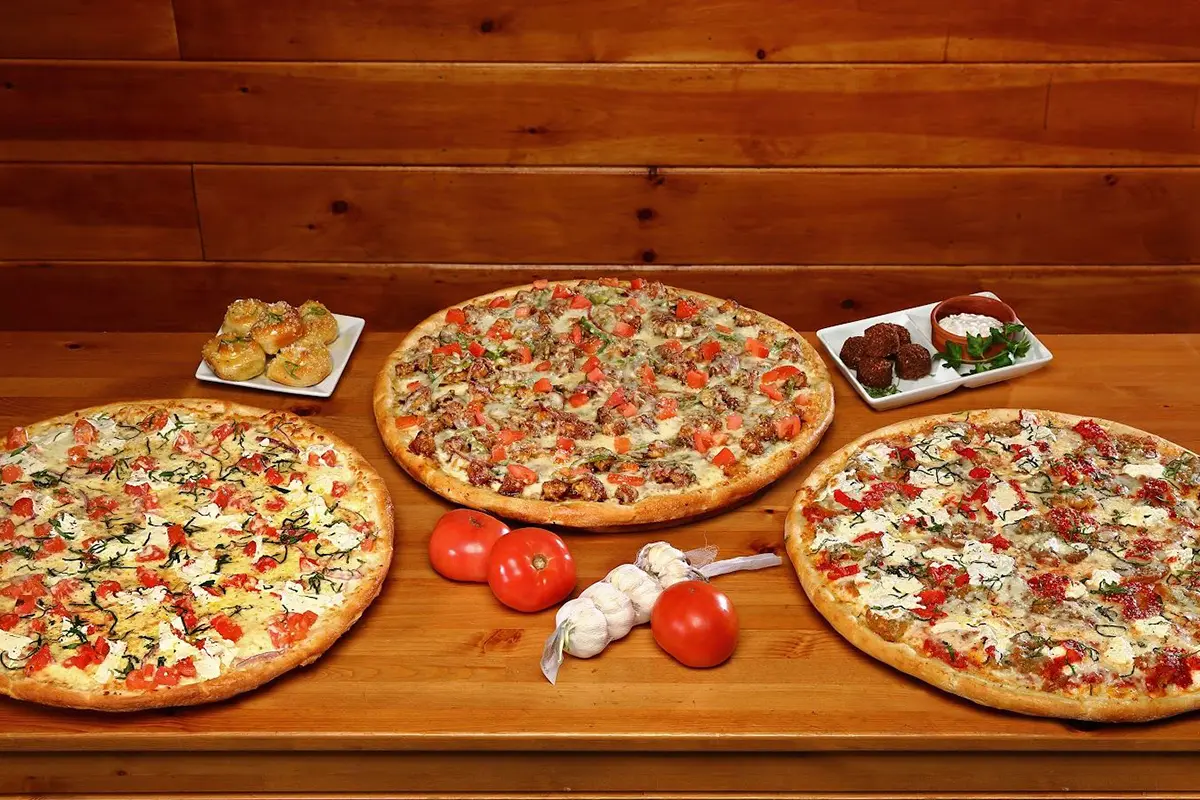 Three pizzas on a wooden table with tomatoes and other food.